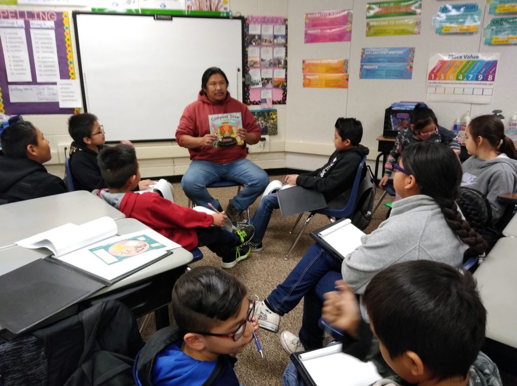 Navajo children sit in a classroom listening to an adult who is reading from a children's book called Compost Stew